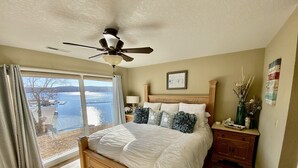 A bedroom with a large bed, a ceiling fan, a nightstand, a dresser, and a view of the water outside.