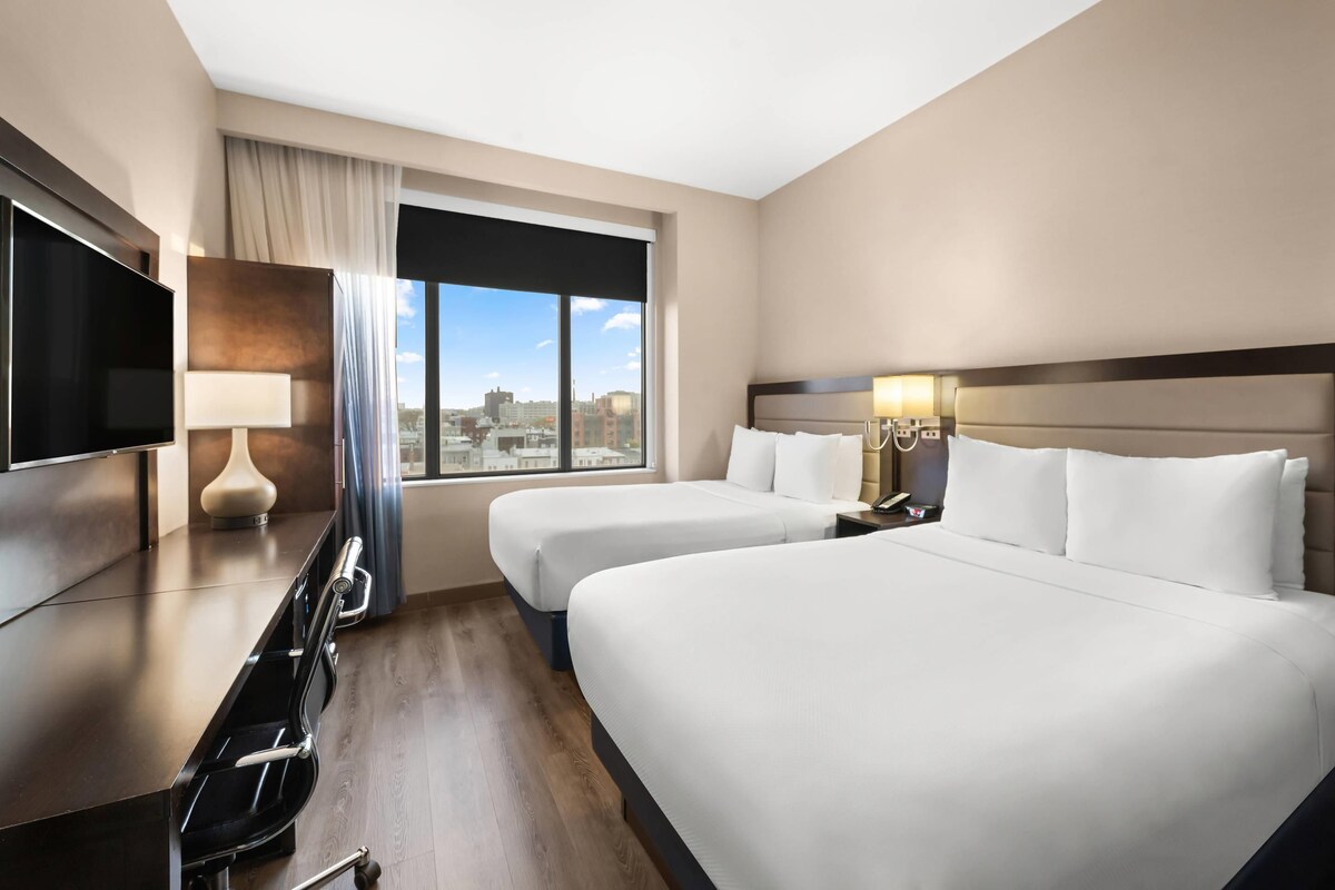 A hotel room with two beds, a TV, a desk, and a window with a city view.
