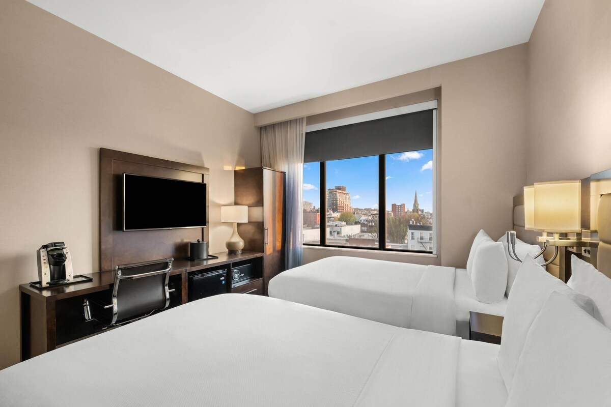 A hotel room with a large bed, a desk with a coffee maker, a TV, and a window with a city view.