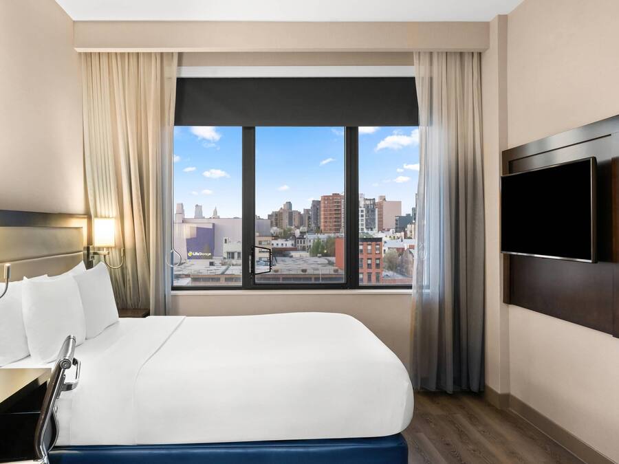 A hotel room with a large bed, a TV mounted on the wall, and a window with a city view.