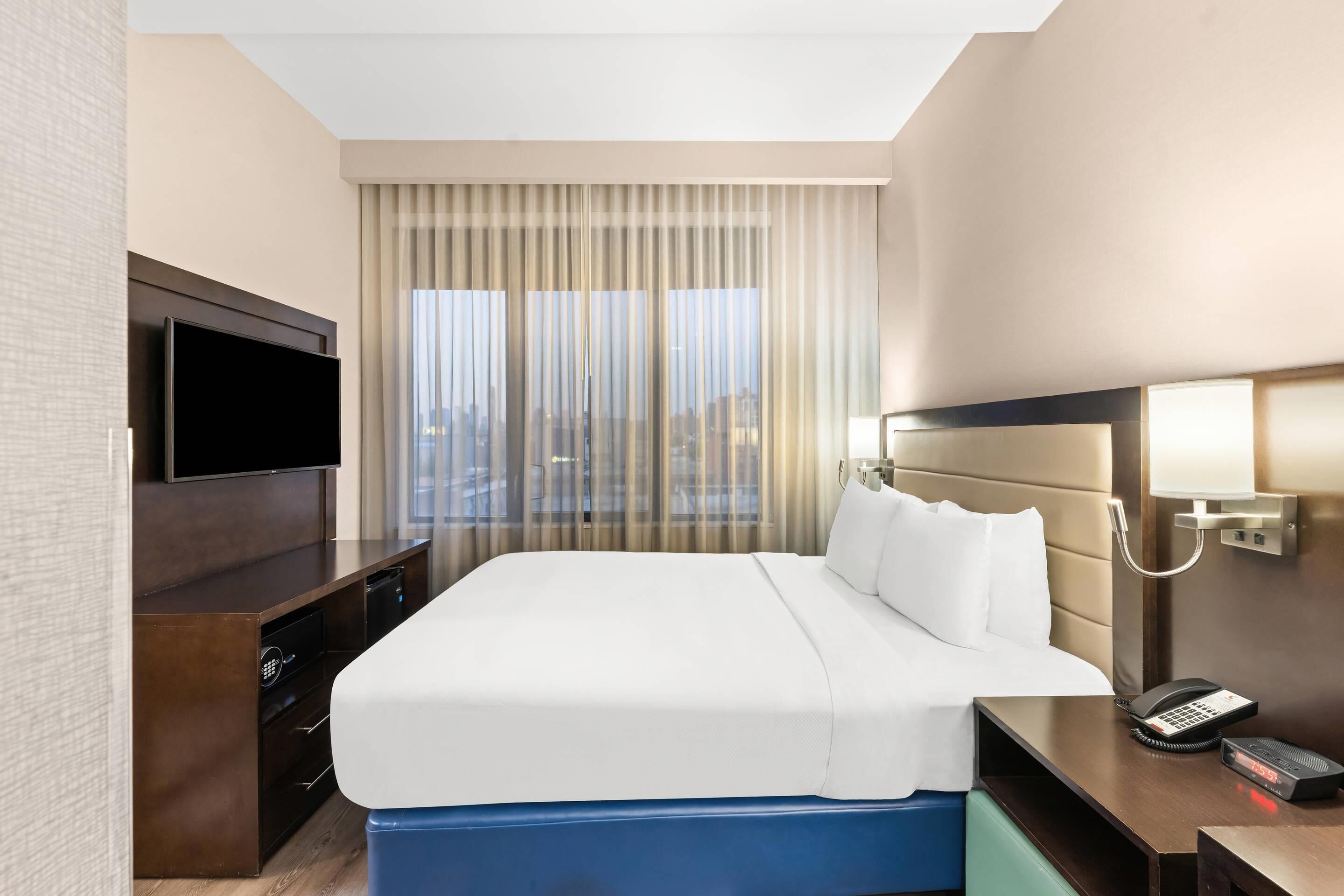 A hotel room with a large bed, a TV on a wooden dresser, a bedside table with a phone, and a view of the city through sheer curtains.