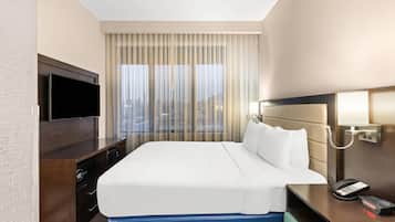 Habitación de hotel con una cama grande, un televisor sobre una cómoda de madera, una mesita de noche con teléfono y vistas a la ciudad a través de cortinas translúcidas.