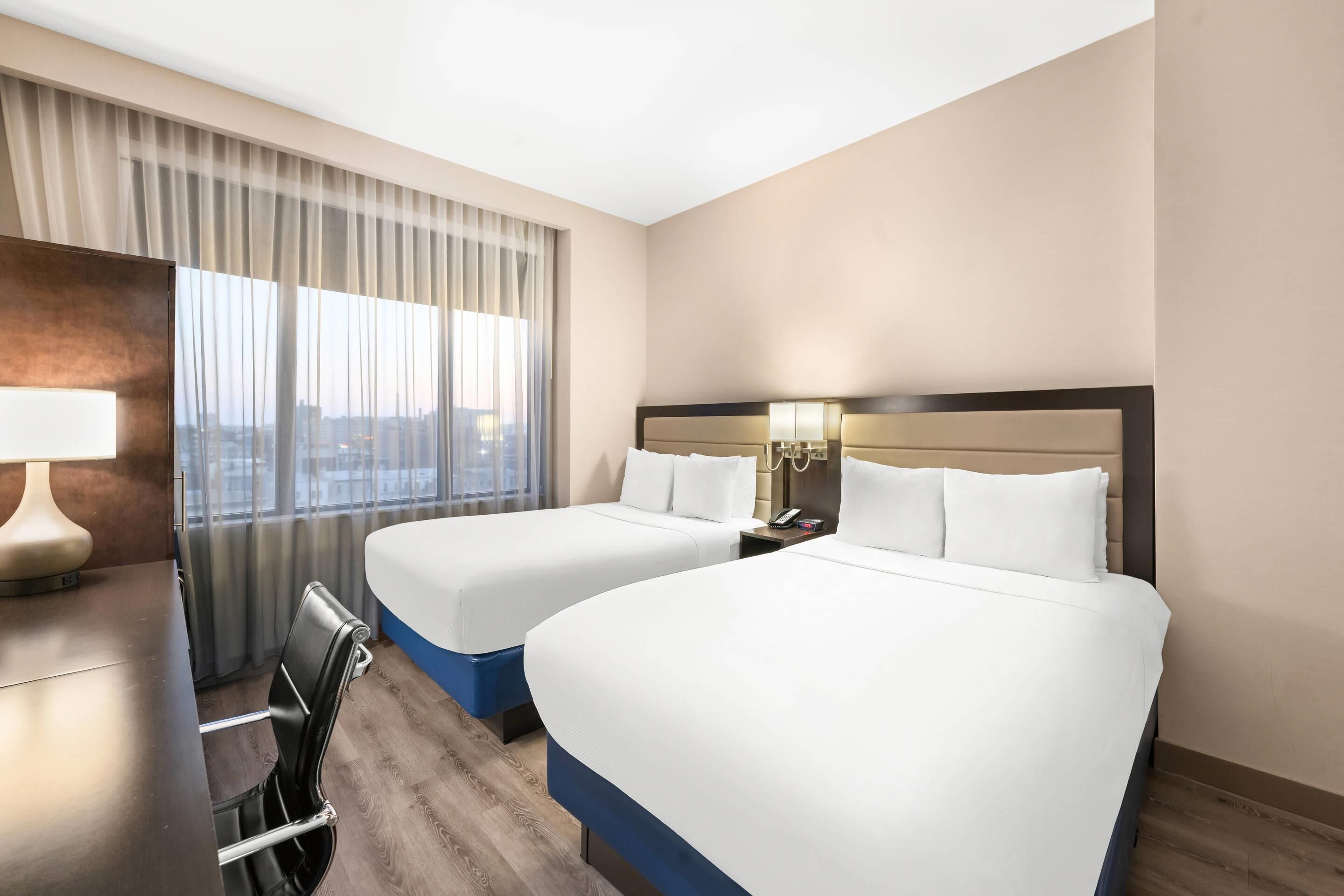 A hotel room with two beds, a desk, a chair, and a view of the city.