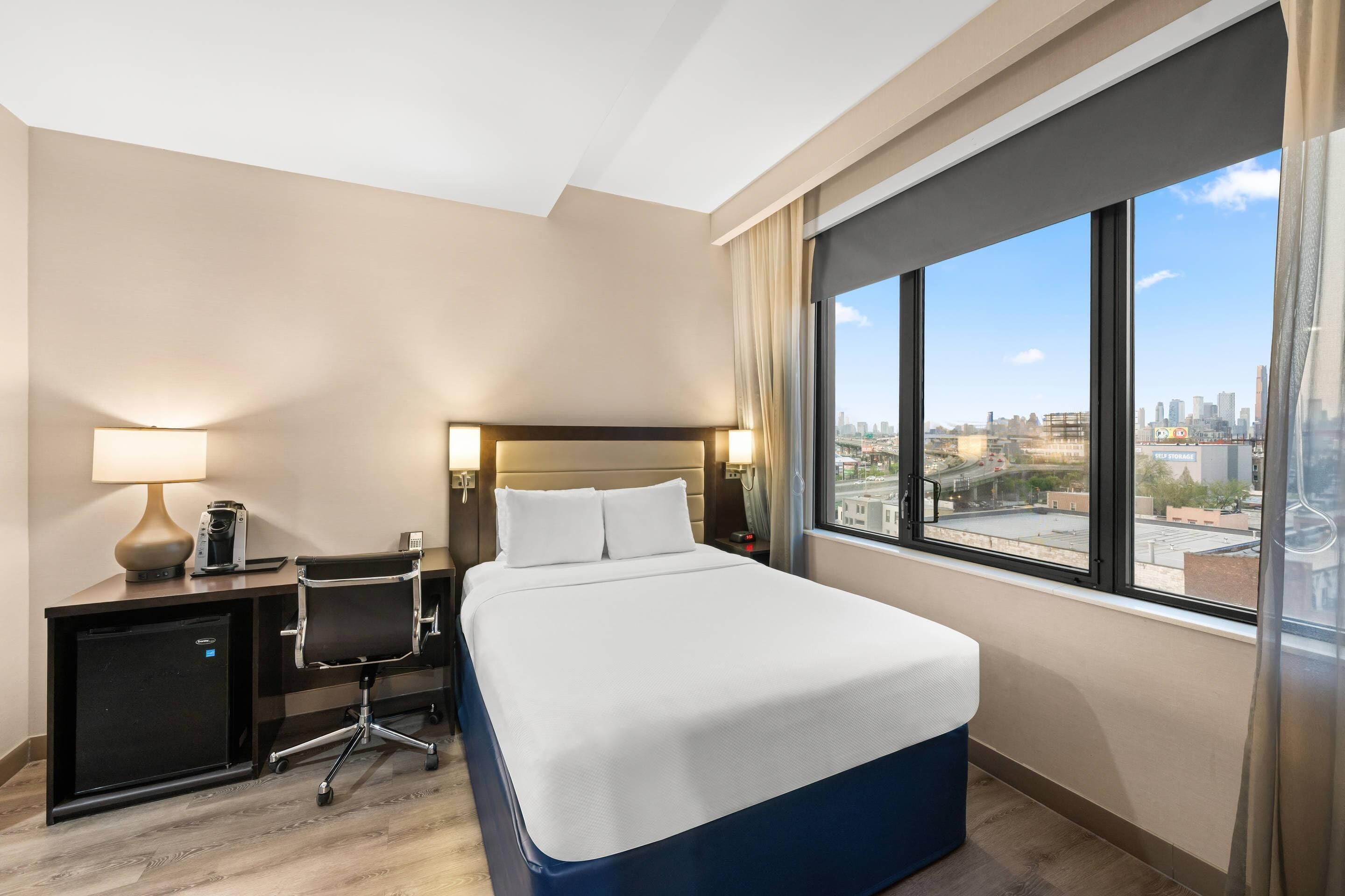 Une chambre d’hôtel équipée d’un lit, d’un bureau, d’une chaise, d’une lampe et d’une grande fenêtre donnant sur la ville.