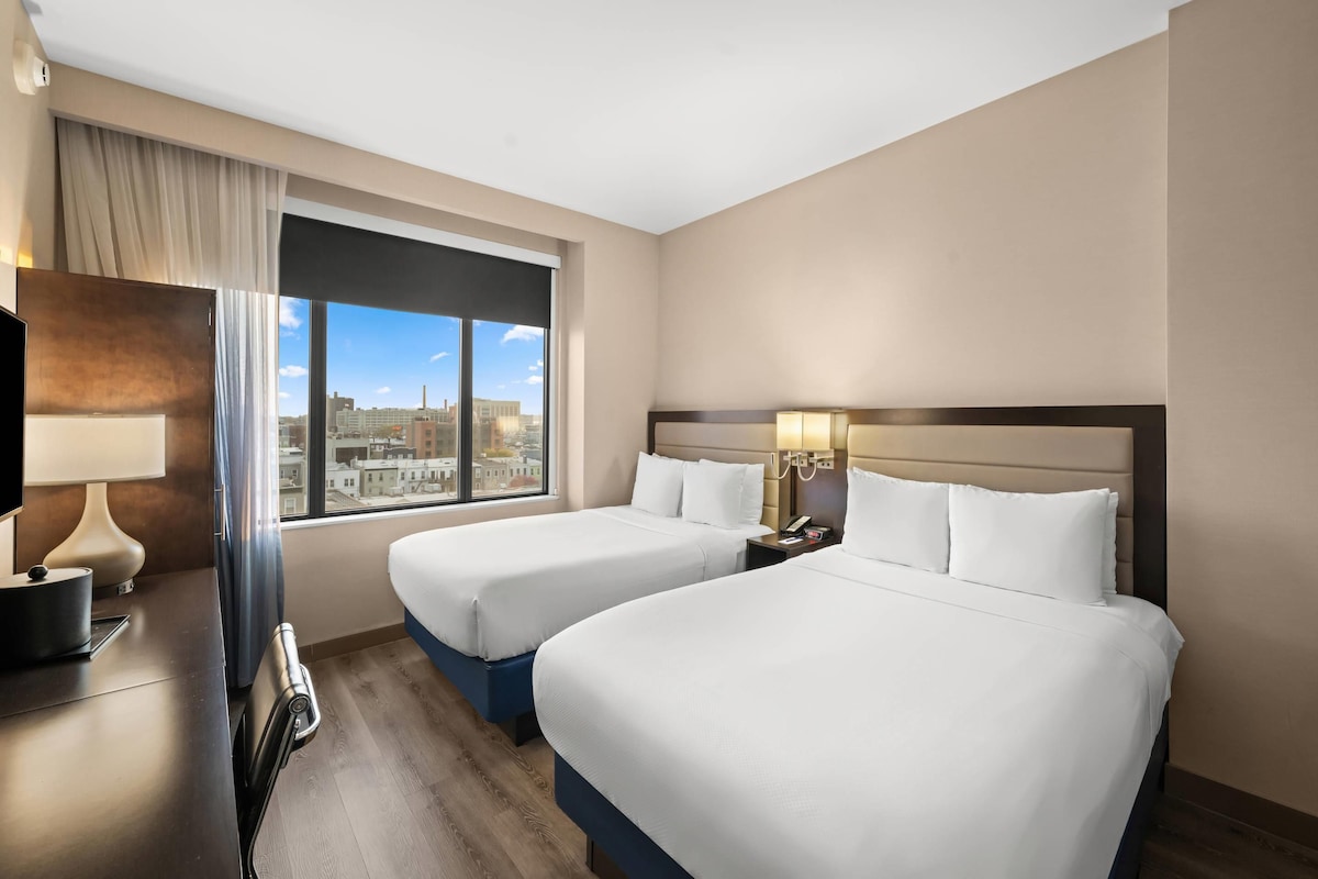 A hotel room with two beds, a desk with a lamp, a chair, a nightstand with a phone, and a window with a city view.
