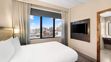 Habitación de hotel con una cama grande, un televisor montado en la pared, un baño con espejo y vista a la ciudad desde la ventana.