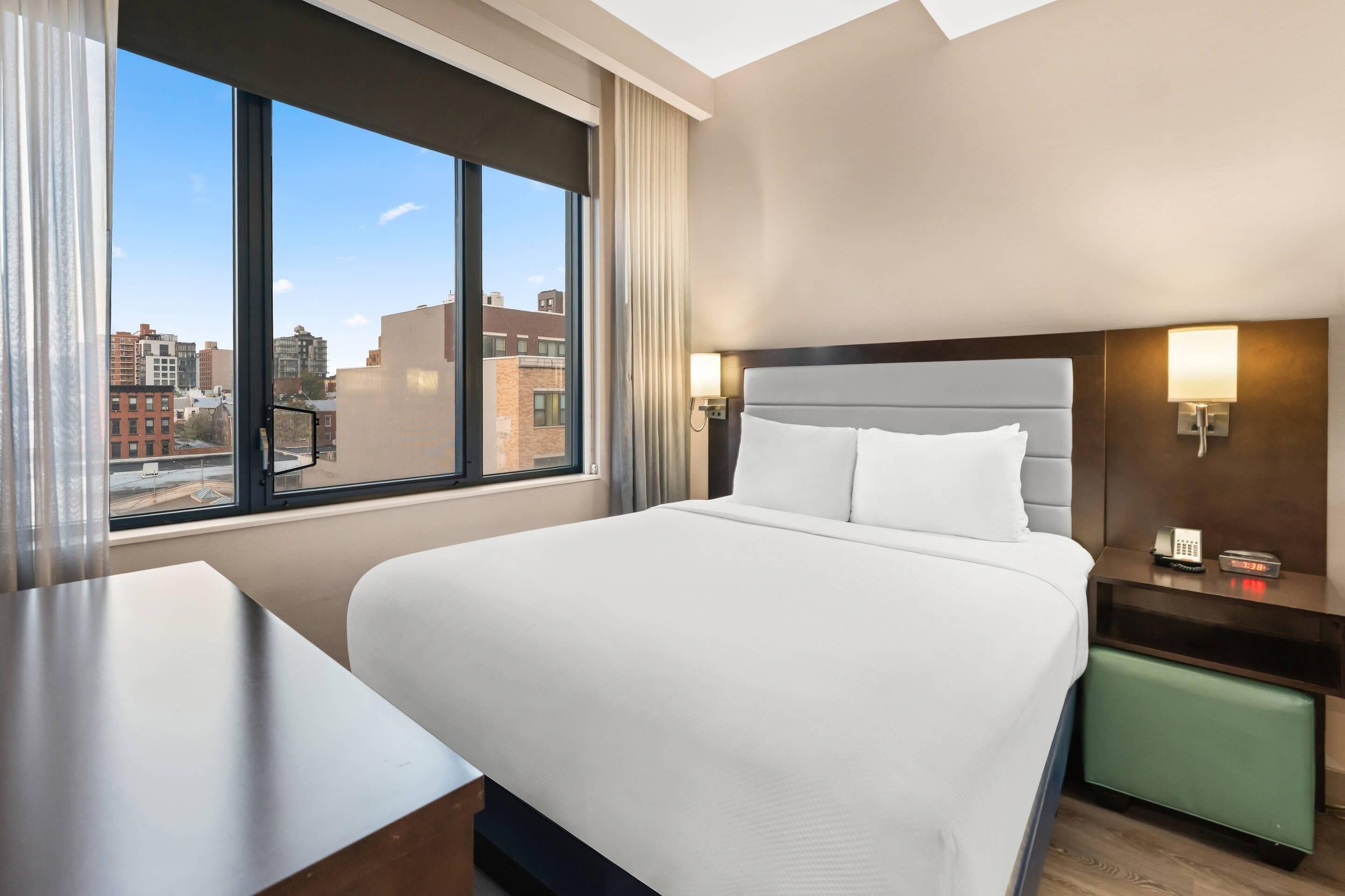 A hotel room with a large bed, a desk, a green chair, a nightstand with a lamp, and a view of the city through large windows.