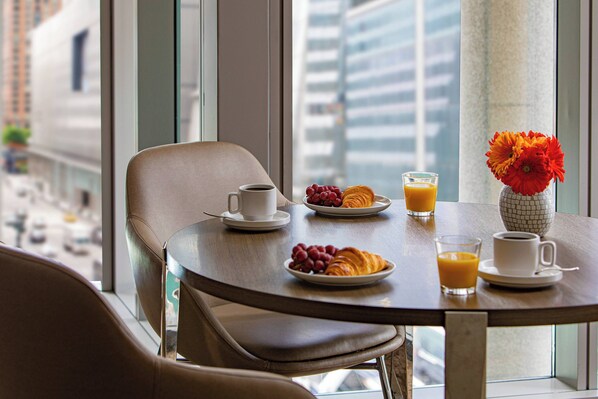 Una mesa de comedor con alimentos para el desayuno, un florero con flores y una vista de la ciudad a través de grandes ventanales.