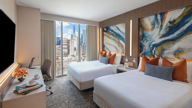 Una habitación de hotel con dos camas, un escritorio con computadora, un televisor y vista a la ciudad.
