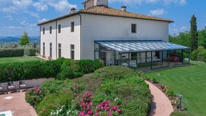 Une maison moderne à deux étages, dotée d’une véranda en verre, entourée de jardins bien entretenus et offrant une vue dégagée sur la campagne.