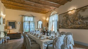 Ein Esszimmer mit einem langen Tisch, gedeckt für eine Mahlzeit, mit rustikalem Ziegelgewölbe, großen Fenstern mit transparenten Vorhängen und einem Gemälde im klassischen Stil an der Wand.