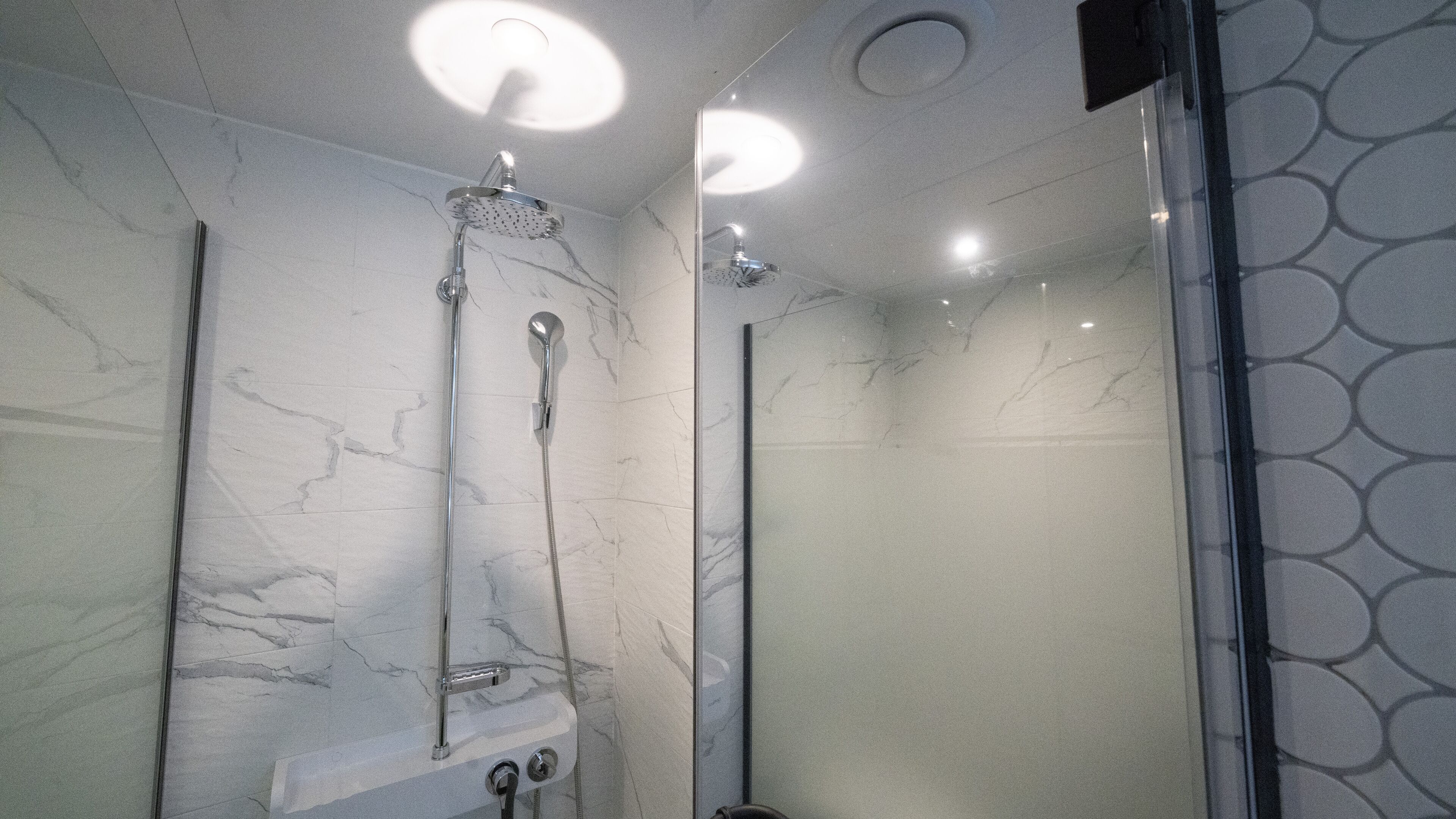 A modern bathroom with a glass shower enclosure, marble walls, and a ceiling light fixture.