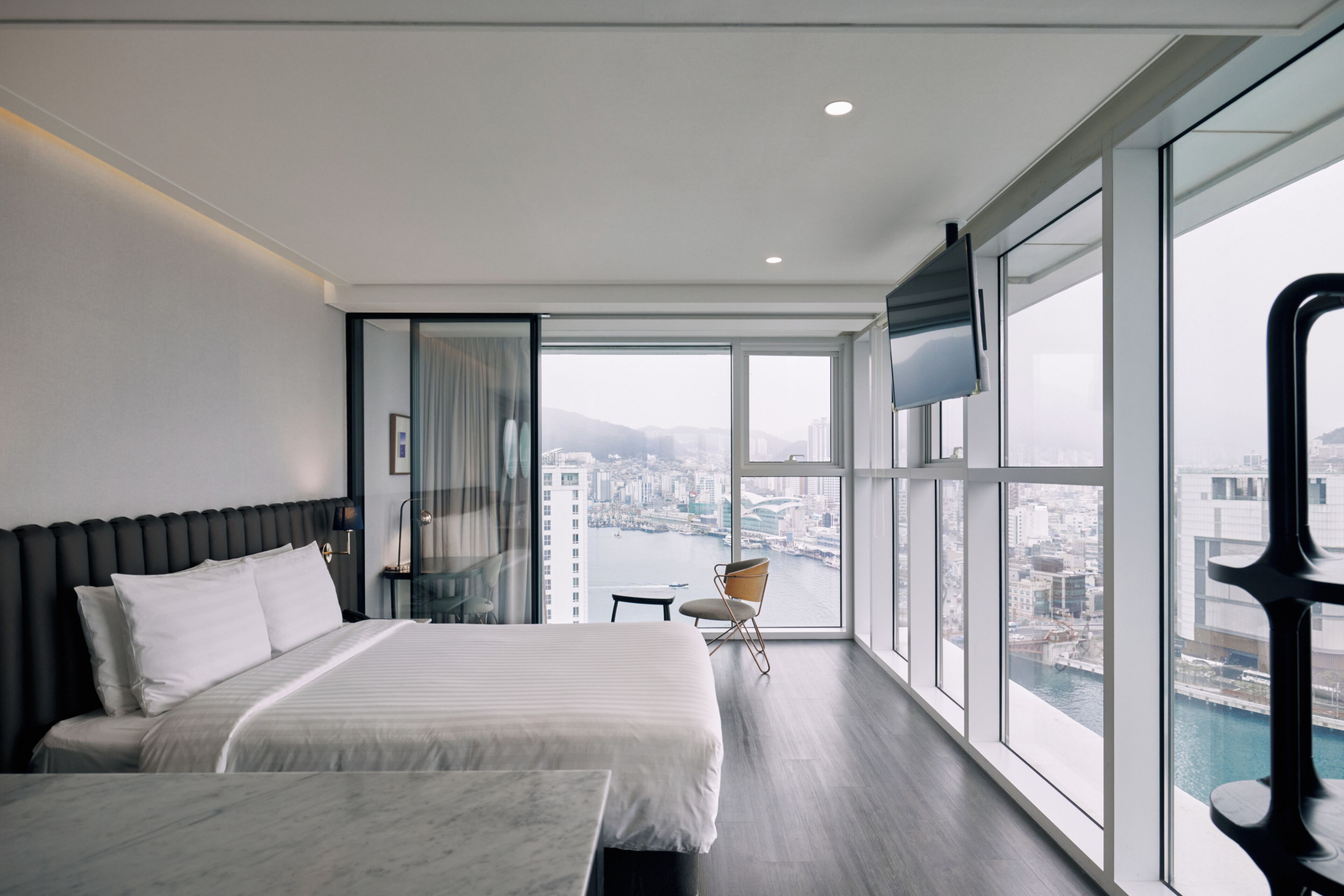 Habitación de hotel moderna con una cama grande, una silla, un televisor y vistas a un paisaje urbano y al agua.