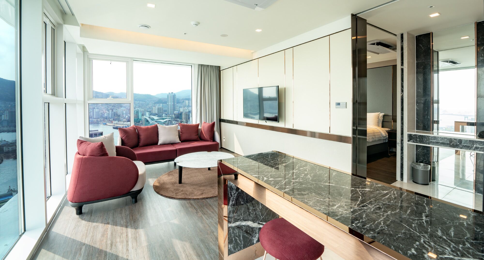 A modern hotel room with a large window offering a city view, a sofa, a round coffee table, and a marble-topped dining table.