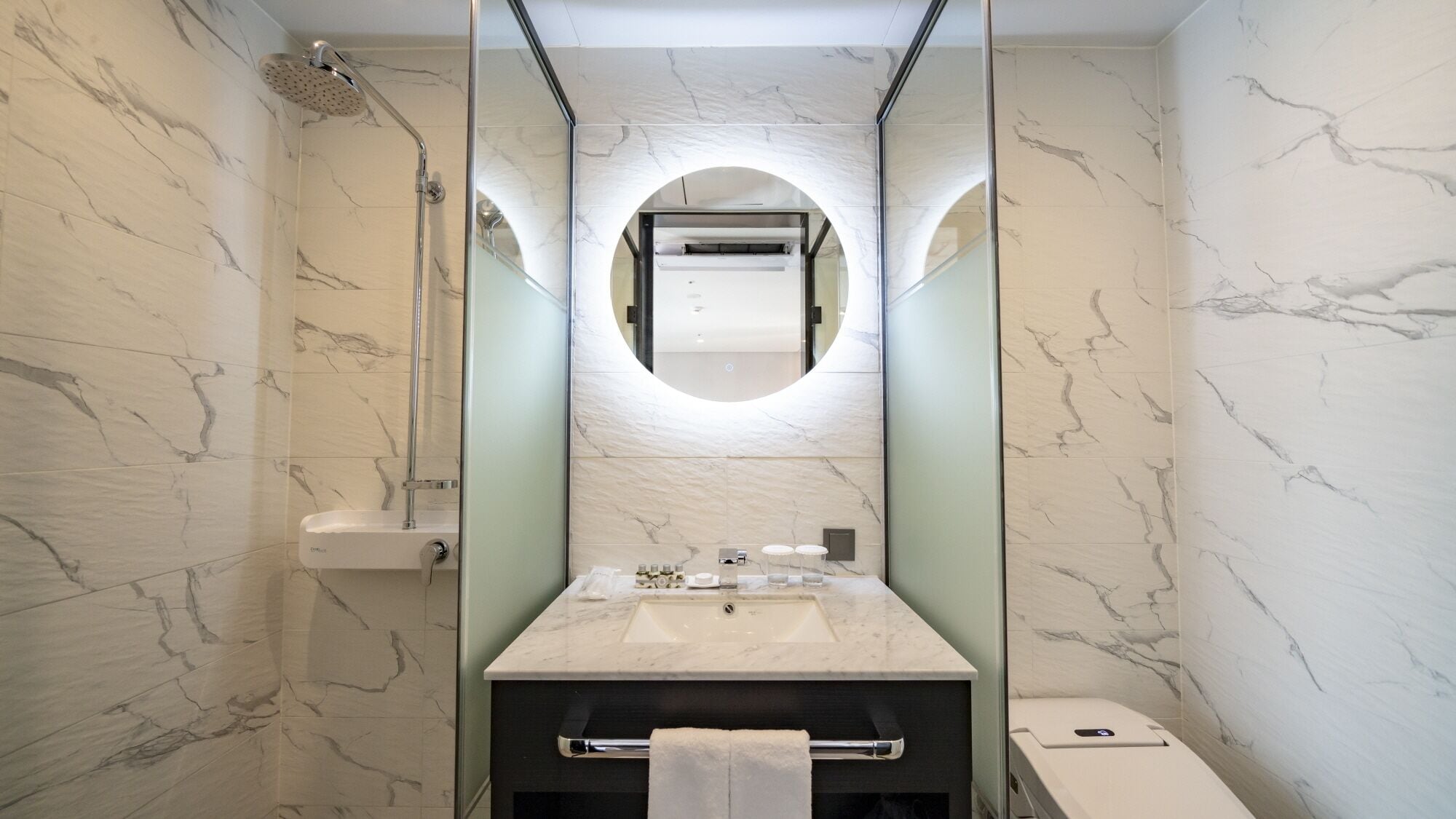 Un baño moderno con tocador de mármol, ducha con cerramiento de vidrio y un espejo grande con marco circular.