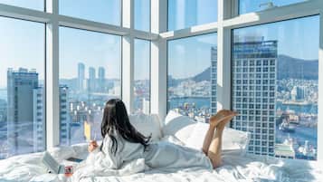 Una mujer recostada en la cama con una computadora portátil y un libro, mirando una vista urbana a través de amplias ventanas.