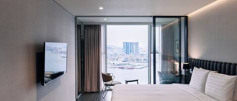 A modern hotel room with a large bed, a TV mounted on the wall, a chair, a small table, and a view of a cityscape through the window.