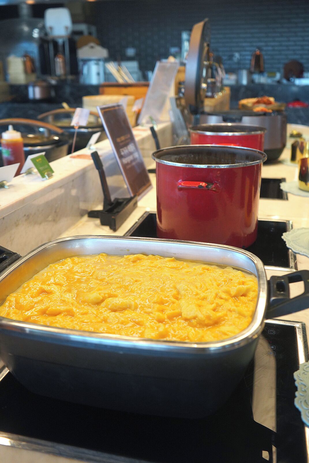 Ein Buffet mit einer großen rechteckigen Servierplatte, gefüllt mit einem gelben Gericht, möglicherweise eine Auflaufform oder etwas Ähnliches, und einem roten Topf im Hintergrund.
