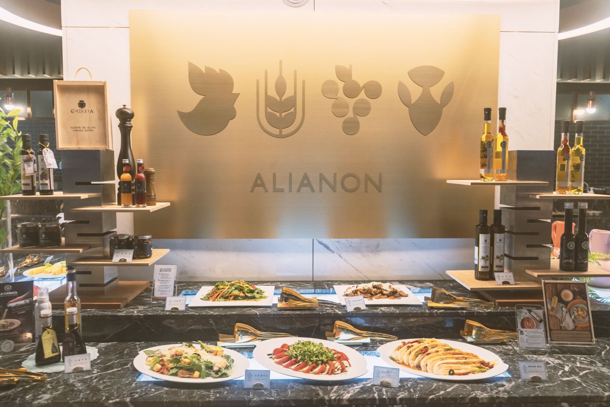 A restaurant counter with various dishes displayed, a wall with the name 'ALIANON', and bottles of wine and olive oil on shelves.