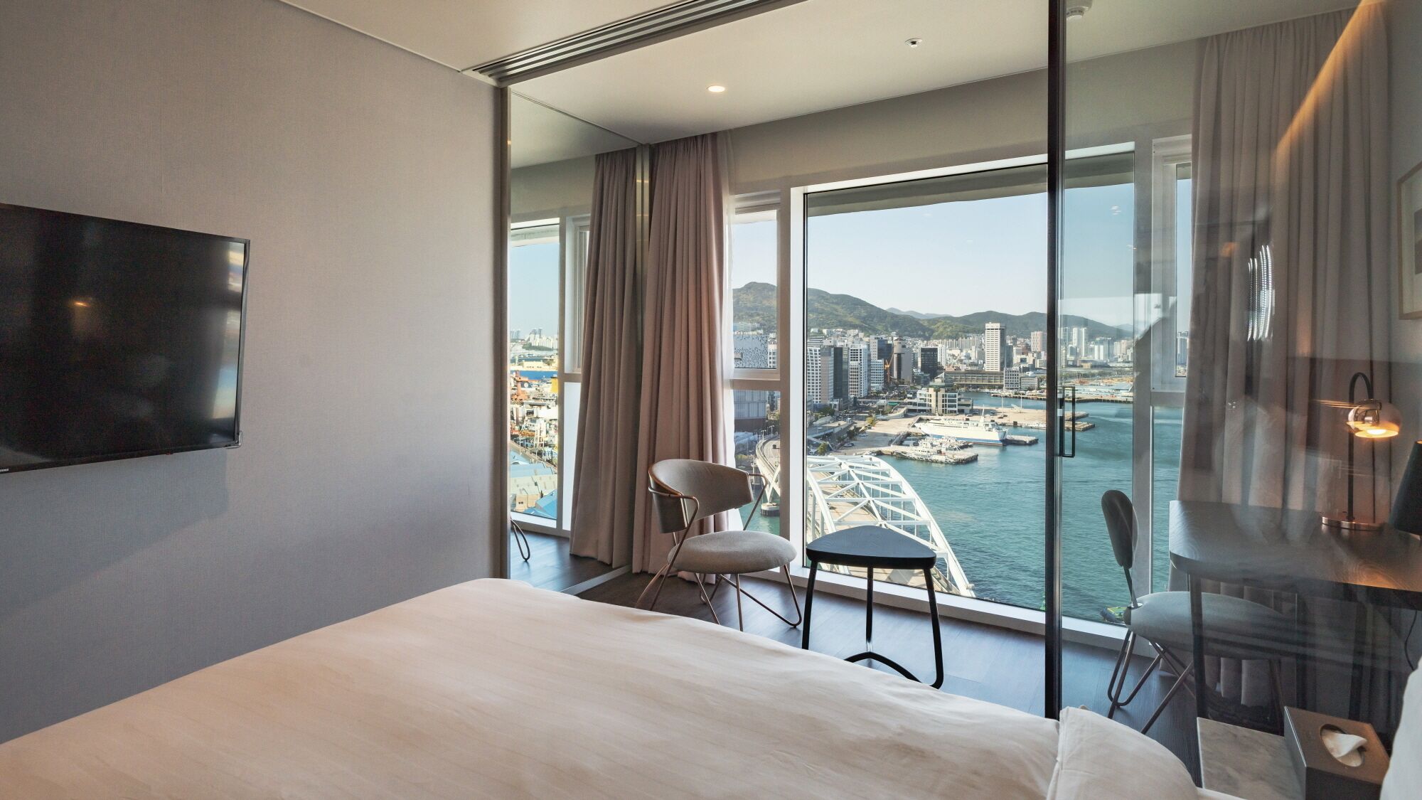 A modern hotel room with a large bed, a TV mounted on the wall, a desk with a chair, and a view of a cityscape and water through floor-to-ceiling windows.