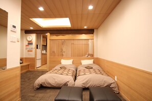 A hotel room with a wooden ceiling, two beds with patterned bedding, a bench, a clock, and a small kitchenette.