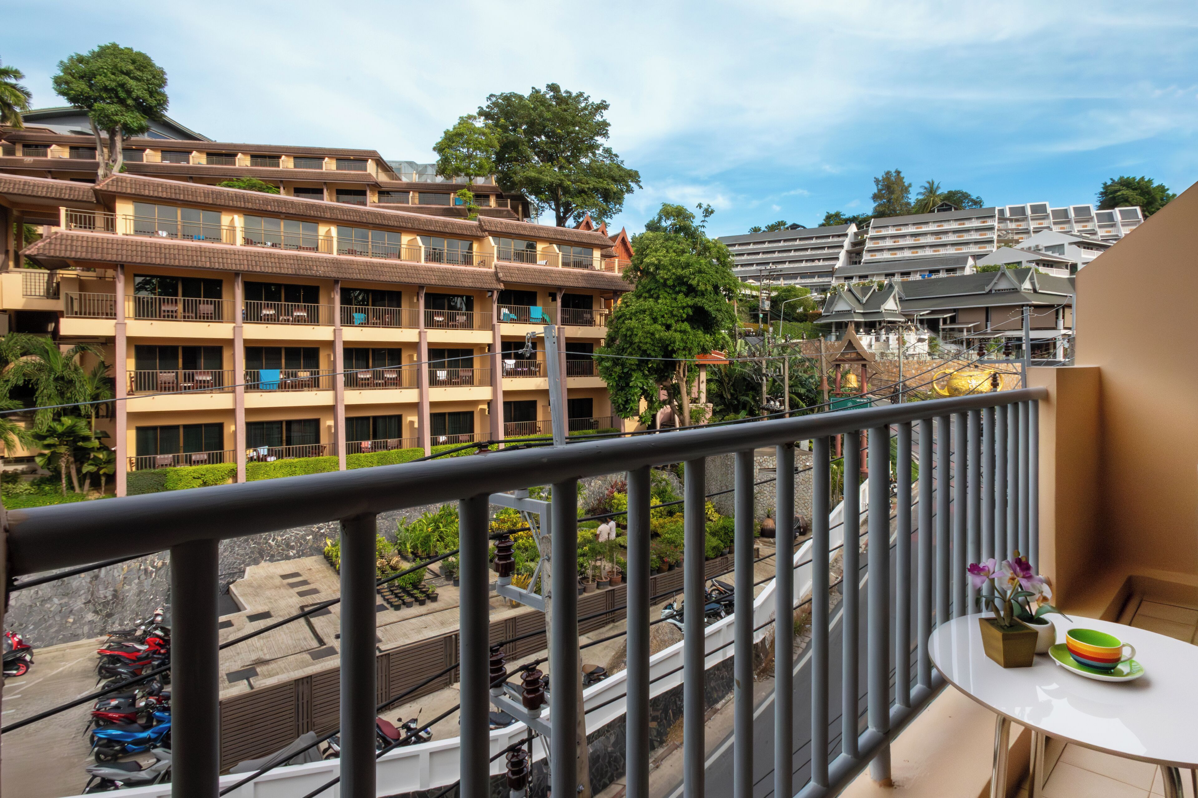 A balcony with a table, potted plants, and a view of a multi-story building with balconies and trees.