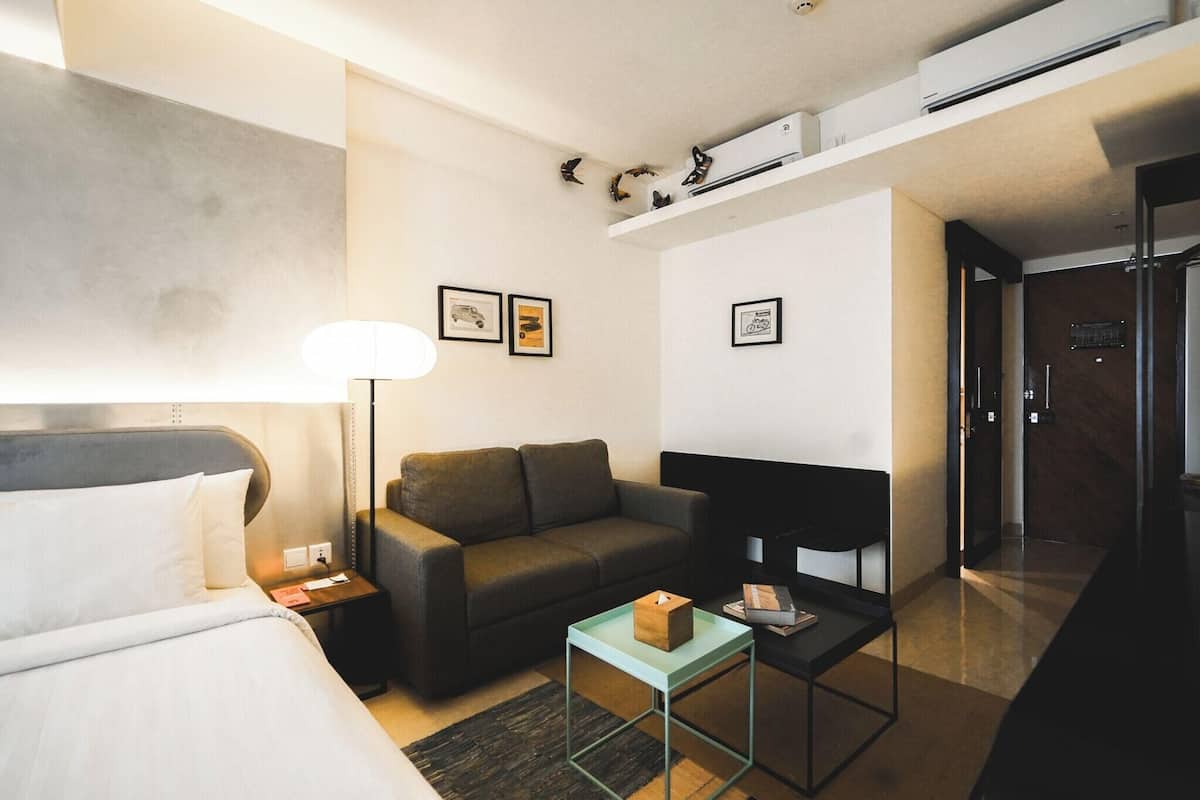 A modern hotel room with a bed, a sofa, a small table, and a desk. There are framed pictures on the wall and a shelf with decorative items.