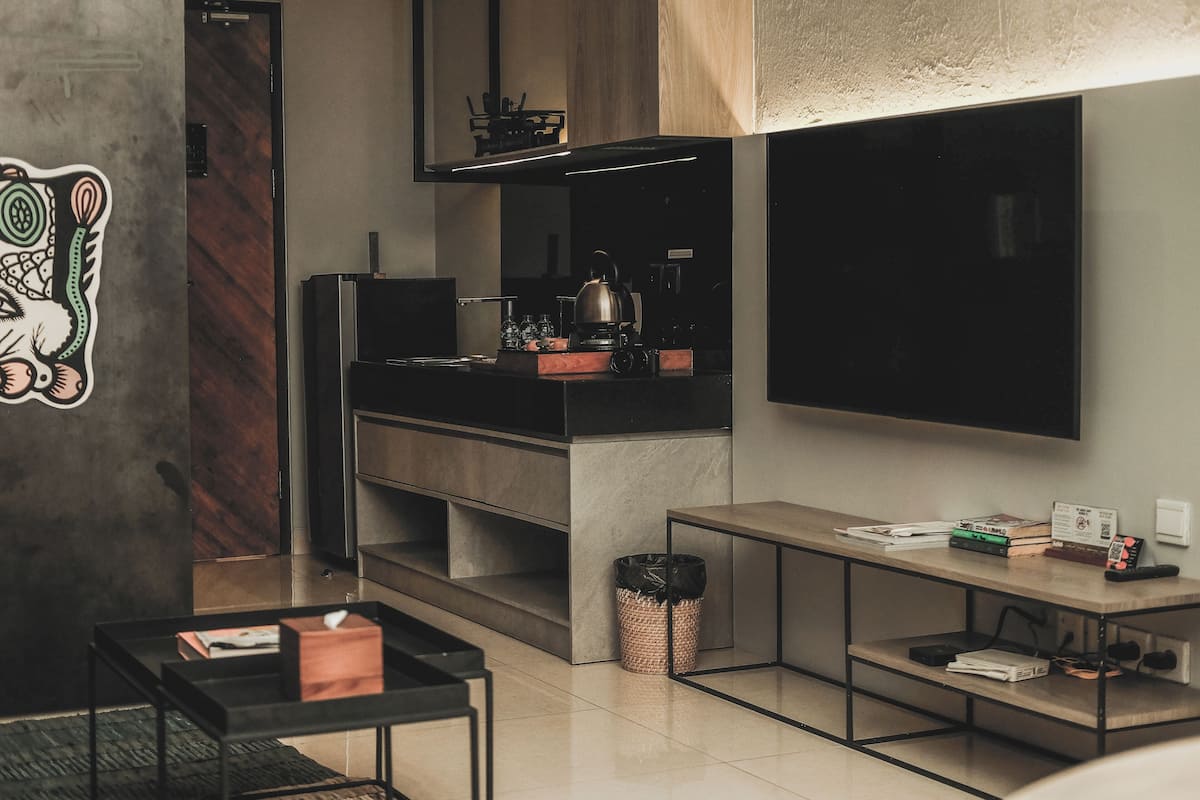 A modern kitchen with a built-in stove, a coffee machine, and a microwave. There is a trash can and a shelf with books and a remote control.