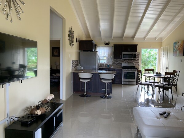 A modern kitchen with dark cabinets, a stainless steel refrigerator, and a microwave. There is a dining area with a white table and chairs.