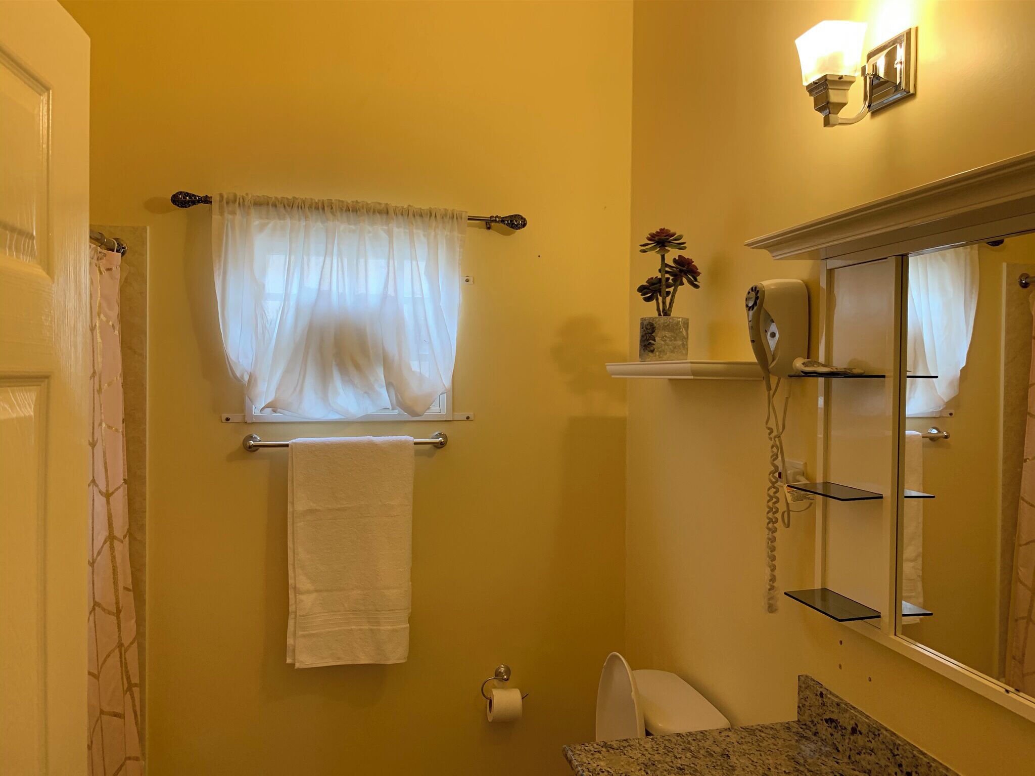 A bathroom with a yellow wall, a towel rack with a white towel, a toilet, a sink with a granite countertop, a mirror, and a wall-mounted phone.