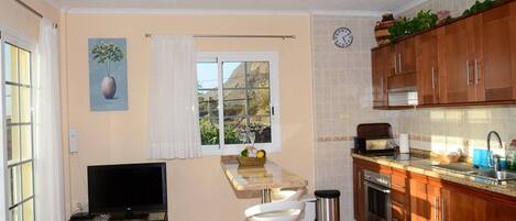 A compact kitchen with a TV, bar stools, and a window with curtains.