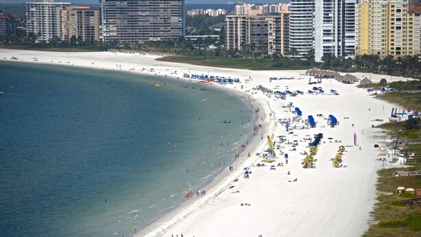 A beach with a white sand shore, a clear blue sea, and several high-rise buildings in the background. - 5th Fl condo, steps to JW Marriott & Beach, Sunset Views, walk to restaurants (Marco Island)
