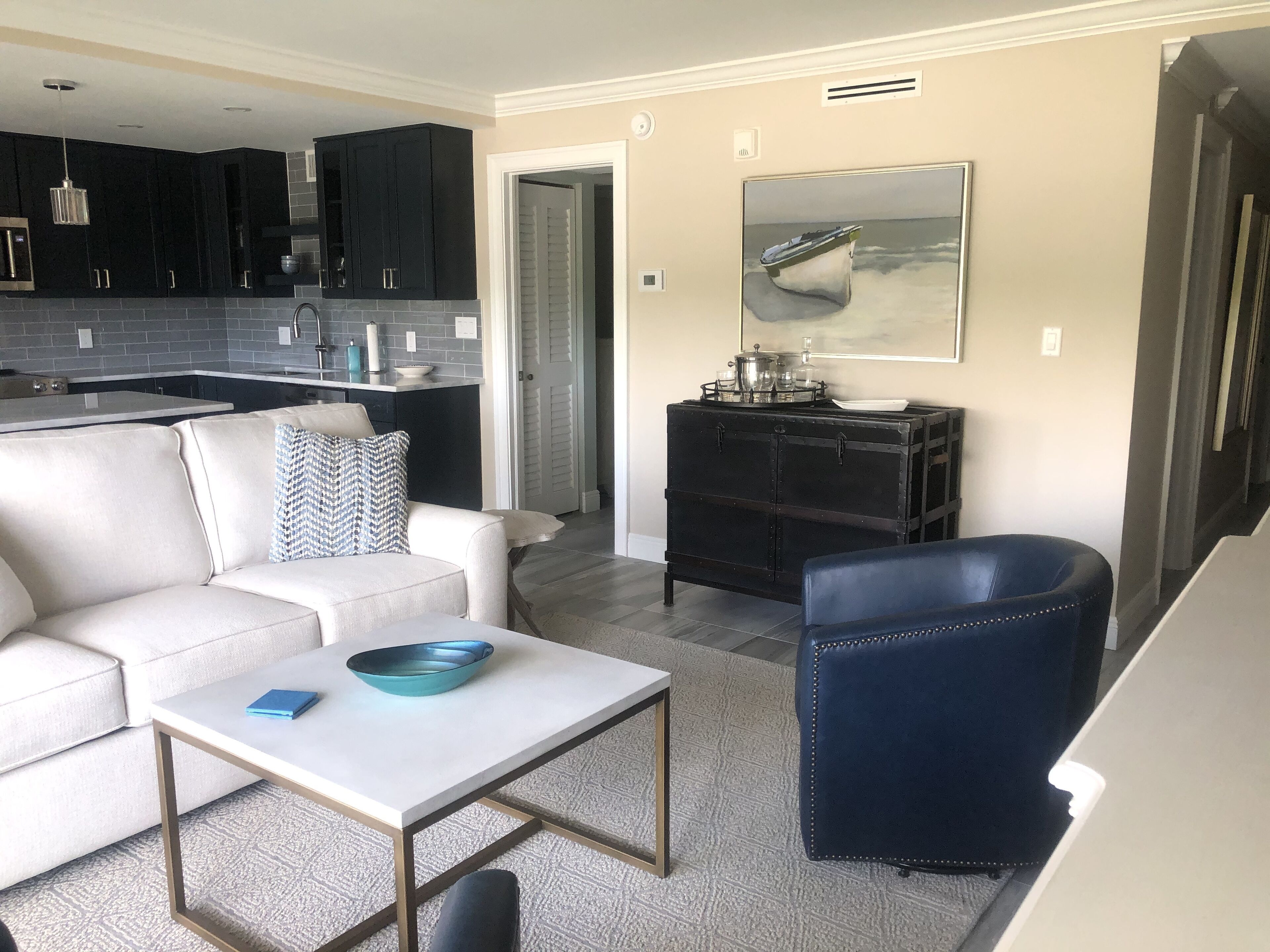 A modern living room with a white sofa, a blue armchair, and a coffee table. A kitchen with dark cabinets and a tiled backsplash is visible in the background.