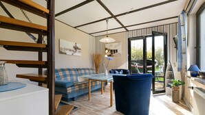 Una sala moderna con sofá, mesa y sillón azul. Se ve una escalera con barandilla de madera, un ventanal grande y una puerta que da al exterior.