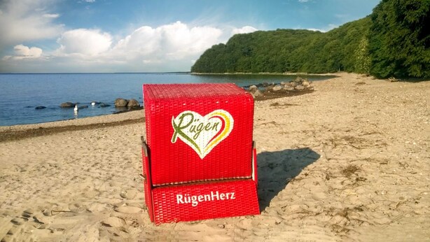 A red box with 'Rügen' and 'RügenHerz' on a beach with a view of the sea and a forested hill.
