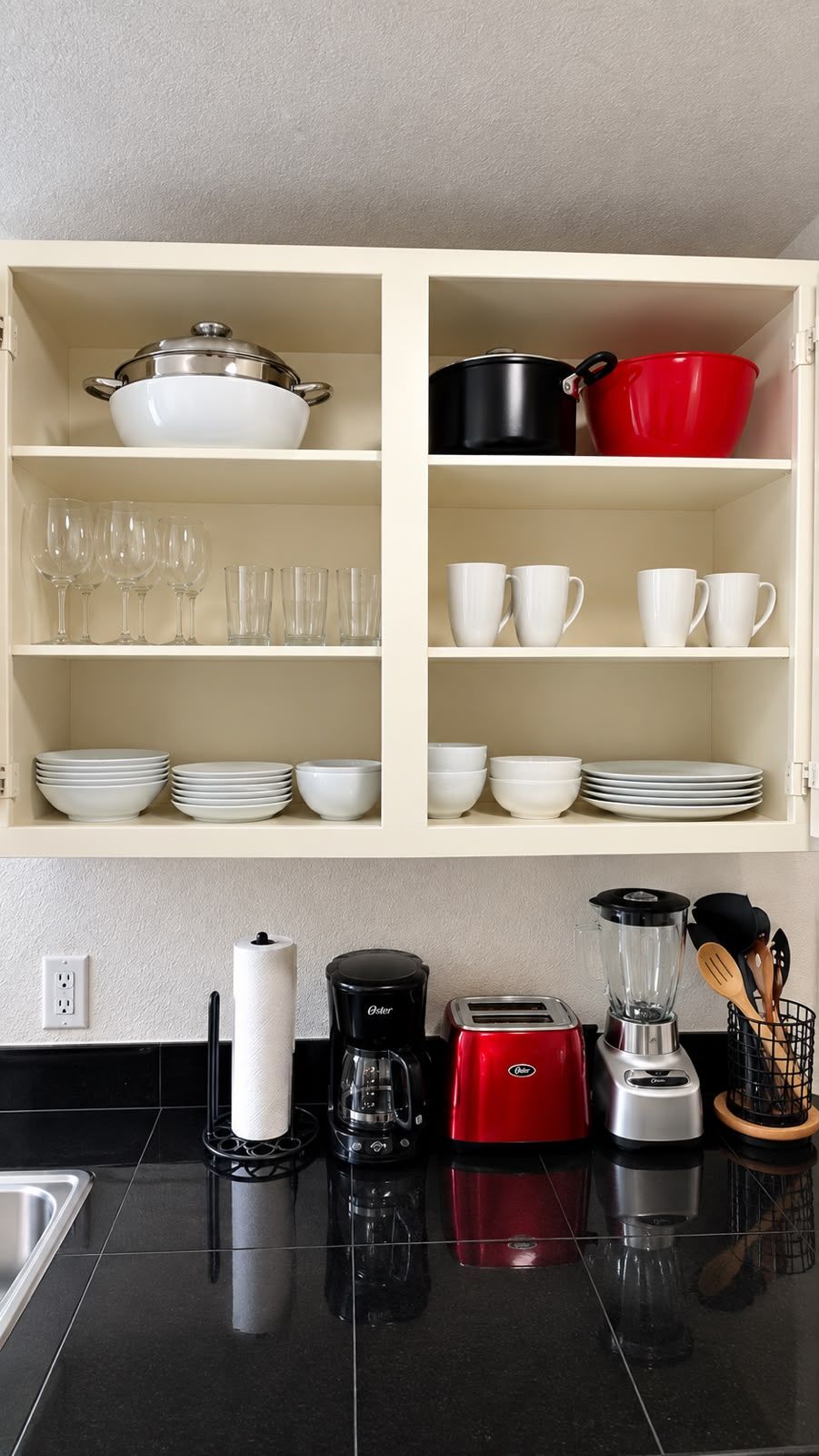A kitchen with a white cabinet containing various dishes and glasses, a black countertop with a coffee maker, toaster, and blender, and a black backsplash.
