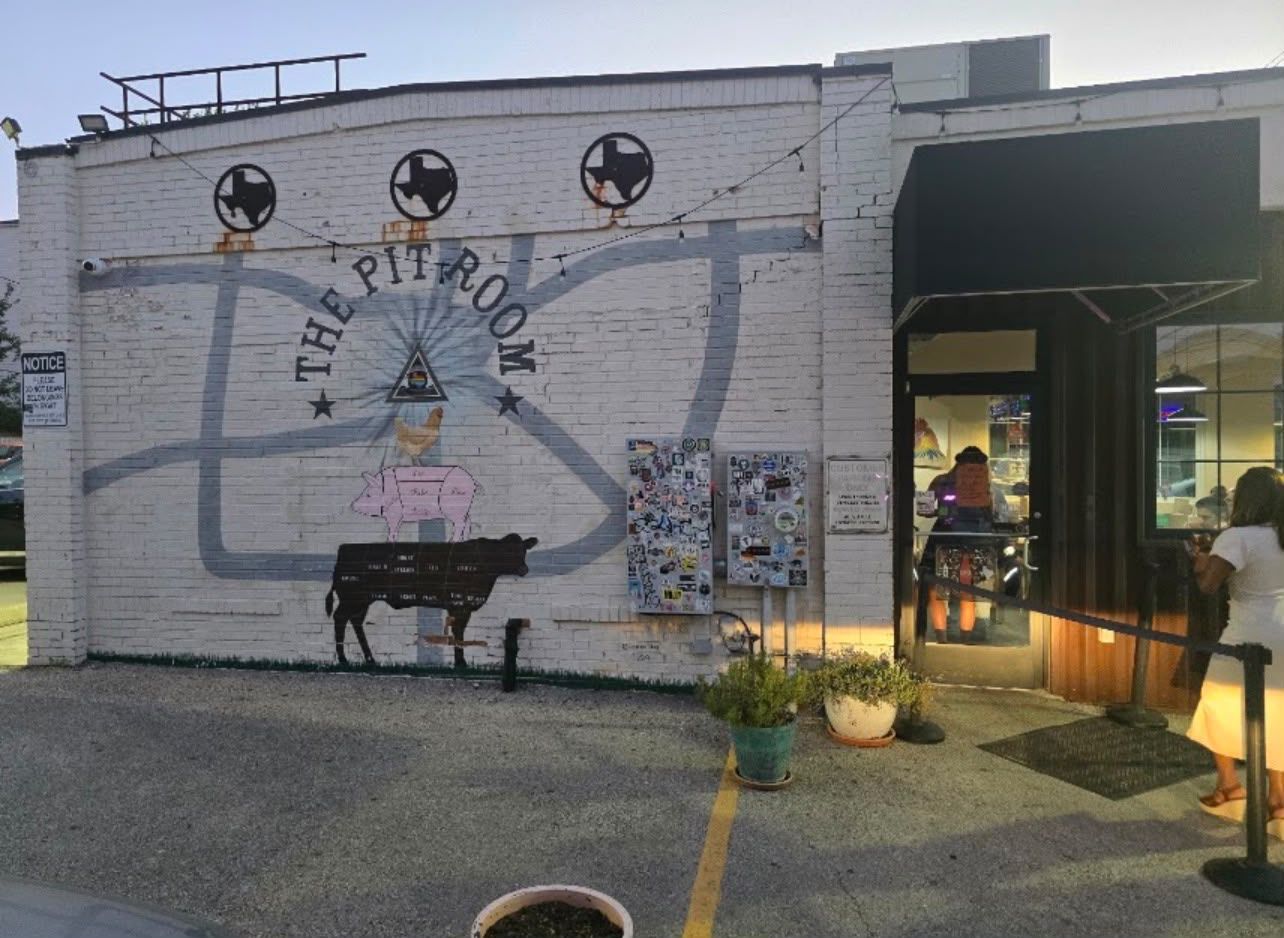 A building with a mural titled 'The Pit Room' featuring a cow and a woman. There are potted plants in front and a person entering the building.