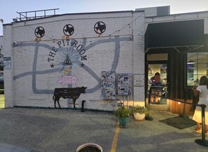 Un edificio con un mural titulado 'The Pit Room' que muestra una vaca y una mujer. Hay plantas en macetas al frente y una persona entrando al edificio.