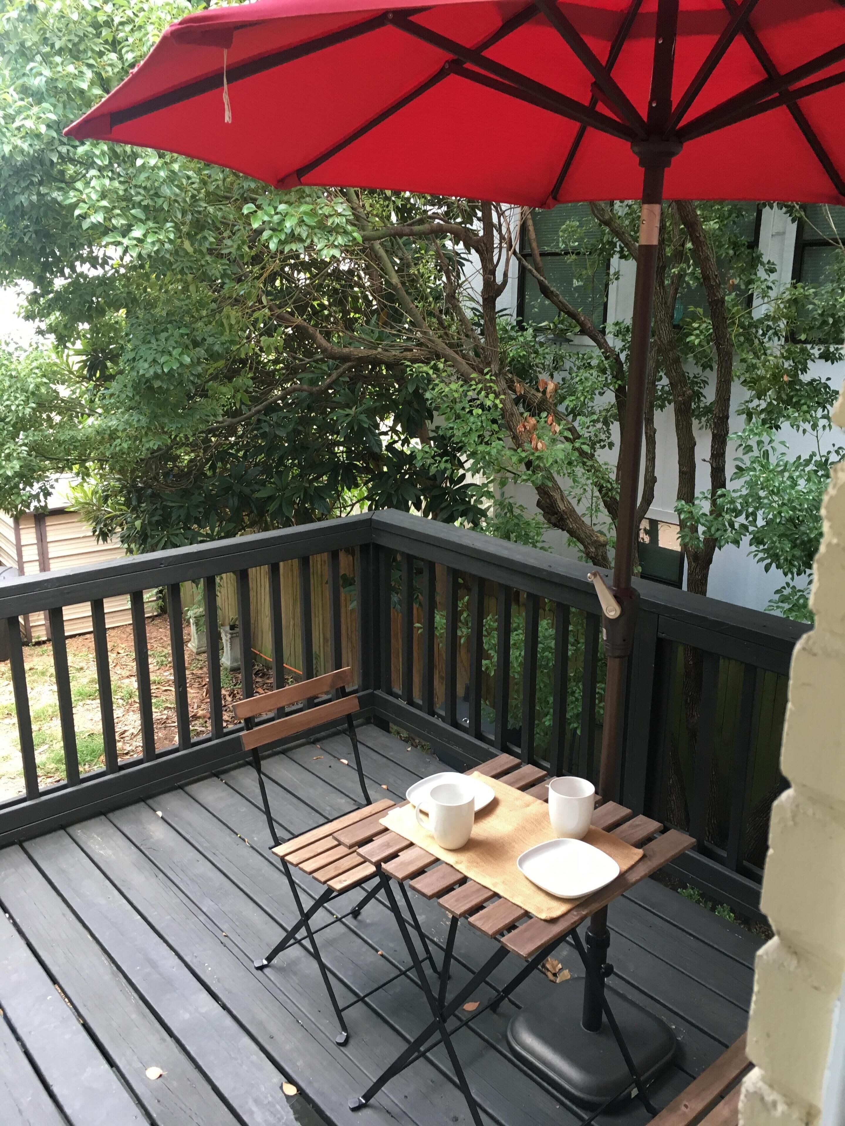 A wooden deck with a table and chairs, a red umbrella, and a white wall in the background.