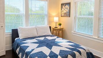 A bedroom with a bed, a ceiling fan, a nightstand with a lamp, and windows with blinds.