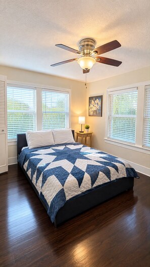 A bedroom with a bed, a ceiling fan, a nightstand with a lamp, and windows with blinds.