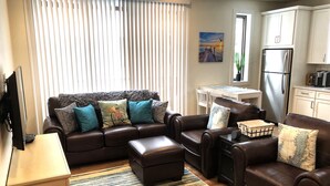 A living room with a brown leather sofa, a coffee table, and a TV stand. There is a kitchen area with a refrigerator and cabinets. - Luxury Downtown Traverse City Condo-Capri 2 (Traverse City)