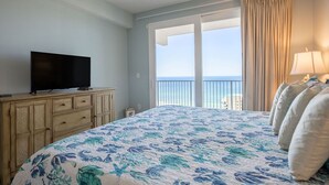 A hotel room with a bed, a television on a wooden dresser, a lamp, and a view of the ocean through a large window. - TOO BEAUTIFUL GULF and SUNSET VIEWS! (Panama City Beach)