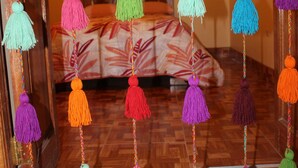 A room with a bed, colorful tassels hanging from the ceiling, and a patterned rug on the floor. - Alpaca Lodge (Chinchero)