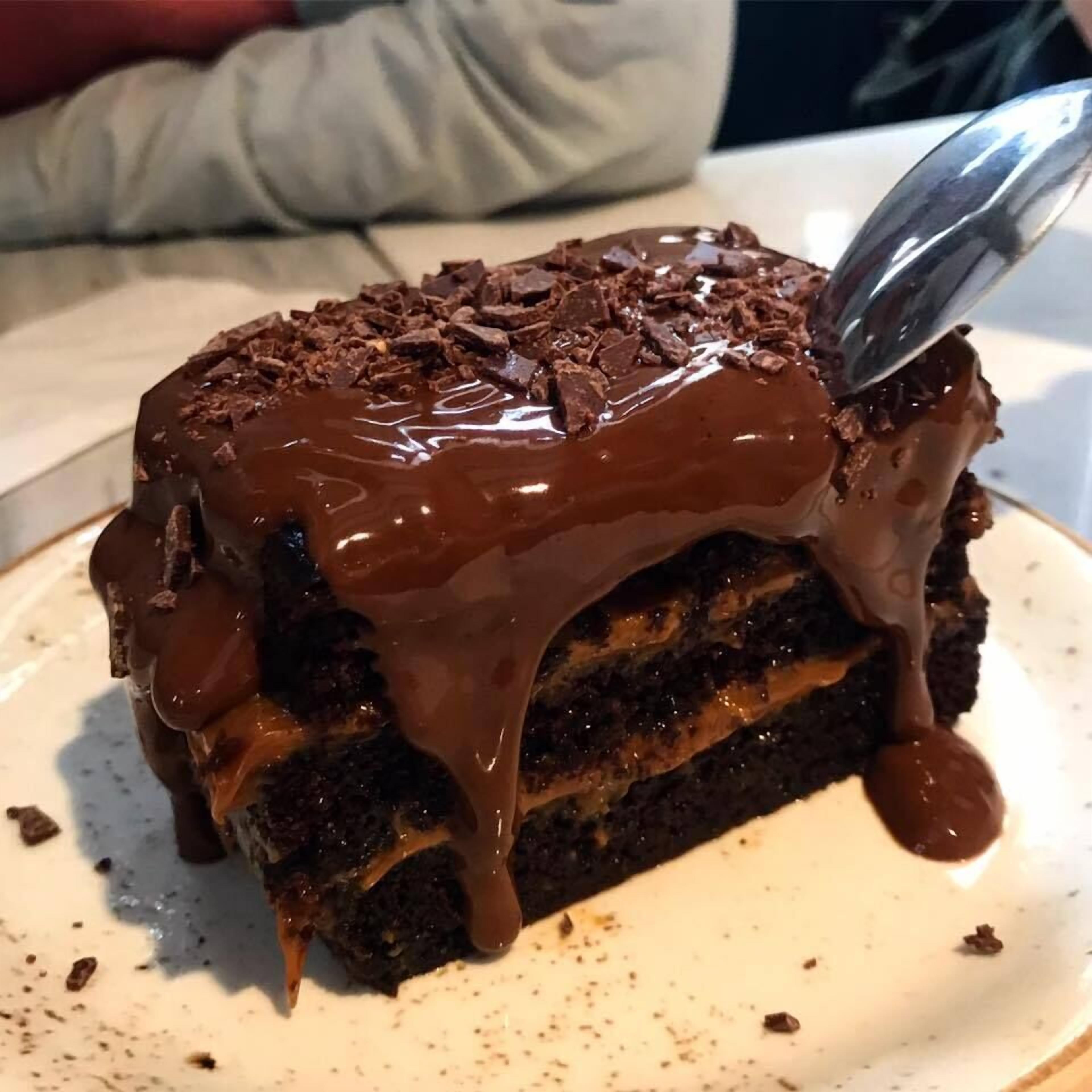 A slice of chocolate cake with a glossy chocolate glaze and chocolate shavings on top, served on a white plate.