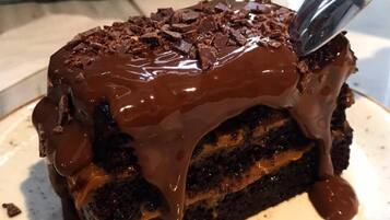 A slice of chocolate cake with a glossy chocolate glaze and chocolate shavings on top, served on a white plate.