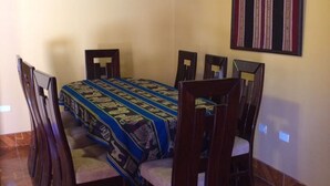A dining area with a round table, six chairs, patterned tablecloth, wooden floor, and a ceiling light fixture. - Alpaca Lodge (Chinchero)