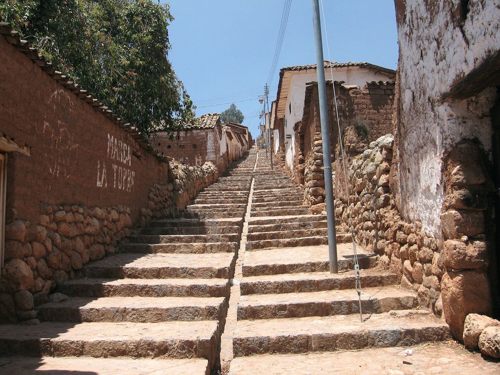 Una estrecha escalera de piedra que asciende por una colina, flanqueada por edificios rústicos con muros de piedra y techos de tejas.
