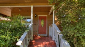 A wooden porch with a red door, white railings, and a small tree to the right.