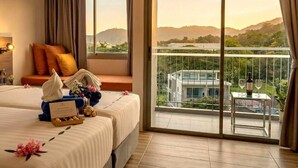 A hotel room with two beds, a balcony with a view of a modern building and hills, and a table with a bottle and glasses.